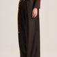 A model wearing dark brown wide-leg pleated trousers with a matching brown belt featuring a tortoiseshell pattern and gold buckle, paired with a brown long-sleeve top and dark loafers, shot against a neutral studio background.