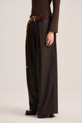 A model wearing dark brown wide-leg pleated trousers with a matching brown belt featuring a tortoiseshell pattern and gold buckle, paired with a brown long-sleeve top and dark loafers, shot against a neutral studio background.