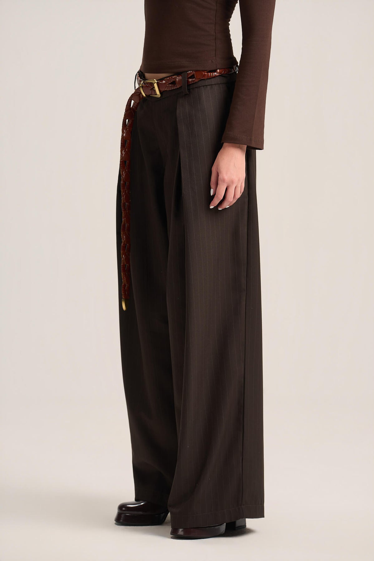 A model wearing dark brown wide-leg pleated trousers with a matching brown belt featuring a tortoiseshell pattern and gold buckle, paired with a brown long-sleeve top and dark loafers, shot against a neutral studio background.