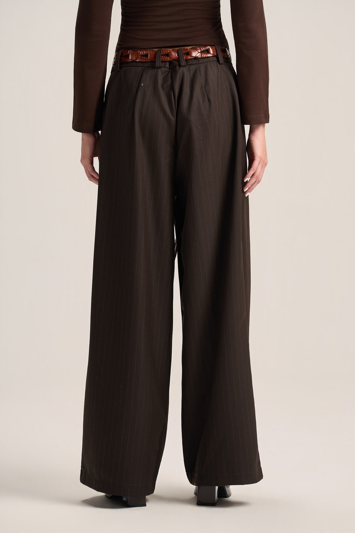 A person shown from the back wearing dark brown wide-leg trousers with a matching dark brown top and a patterned brown belt, standing against a plain light beige studio background.