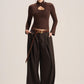 A model stands against a plain light beige studio background wearing a dark brown long-sleeve mock-neck top with a chest cutout, paired with matching dark brown wide-leg trousers and a brown belt.