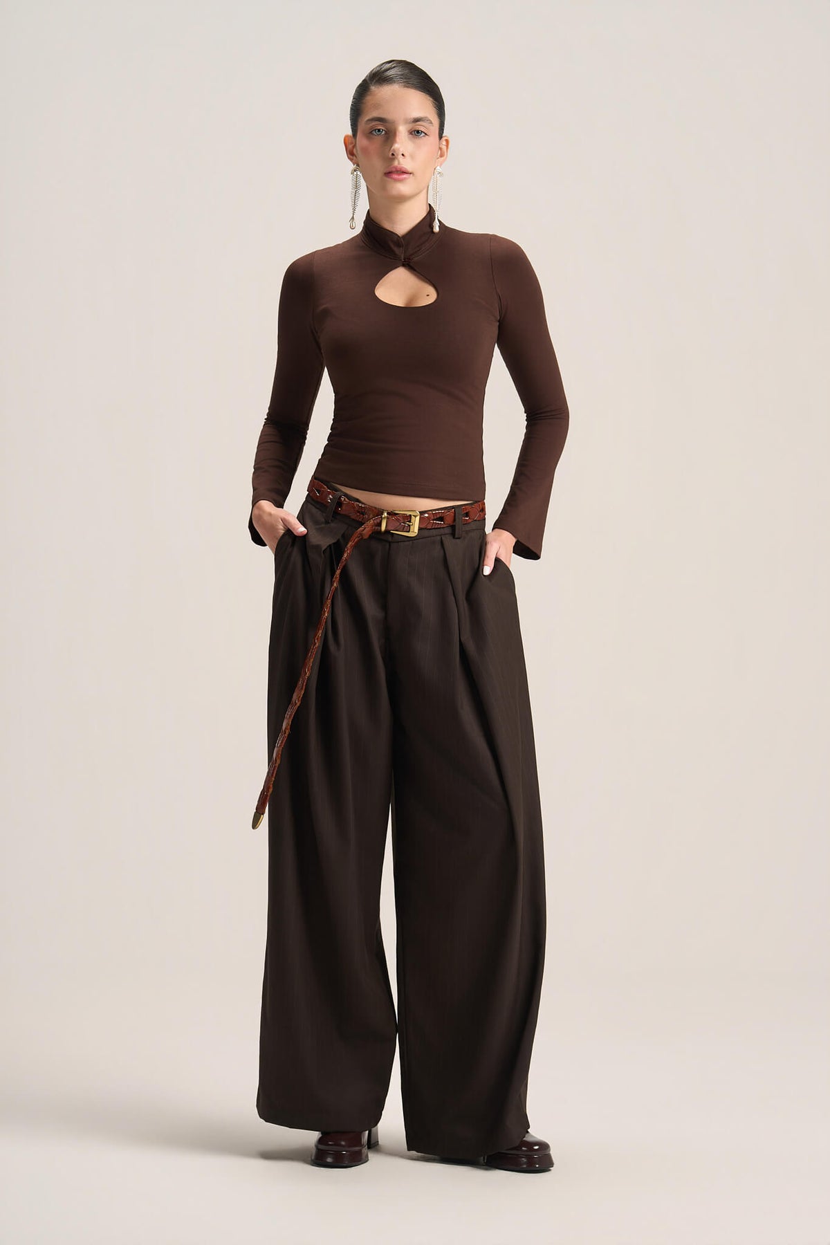 A model stands against a plain light beige studio background wearing a dark brown long-sleeve mock-neck top with a chest cutout, paired with matching dark brown wide-leg trousers and a brown belt.
