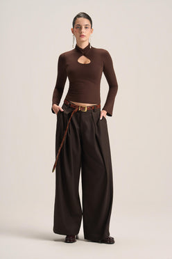 A model stands against a plain light beige studio background wearing a dark brown long-sleeve mock-neck top with a chest cutout, paired with matching dark brown wide-leg trousers and a brown belt.