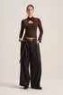 A model stands against a plain light beige studio background wearing a dark brown long-sleeve mock-neck top with a chest cutout, paired with matching dark brown wide-leg trousers and a brown belt.