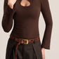 A woman wearing a fitted long-sleeve brown mock-neck top with a keyhole cutout at the chest, paired with dark pleated trousers and a brown belt, against a plain light beige studio background.