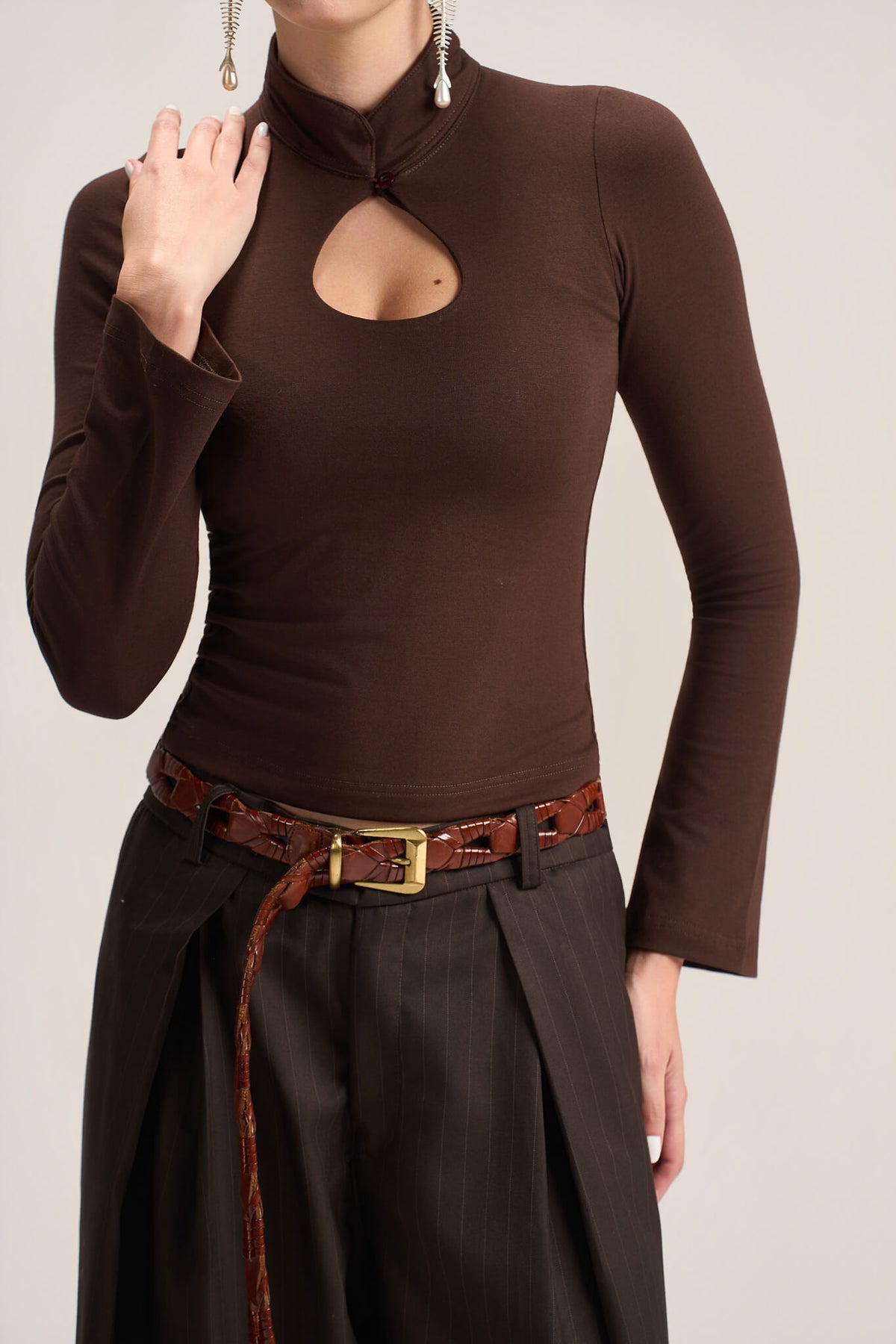 A woman wearing a fitted long-sleeve brown mock-neck top with a keyhole cutout at the chest, paired with dark pleated trousers and a brown belt, against a plain light beige studio background.