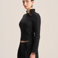 A model wearing a black long-sleeve mock-neck crop top with lace-up detailing at the collar, paired with black wide-leg trousers, against a solid light beige background.