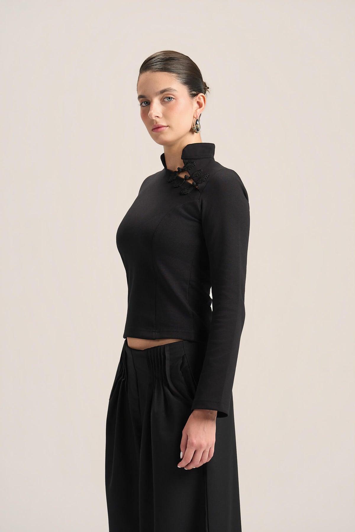 A model wearing a black long-sleeve mock-neck crop top with lace-up detailing at the collar, paired with black wide-leg trousers, against a solid light beige background.