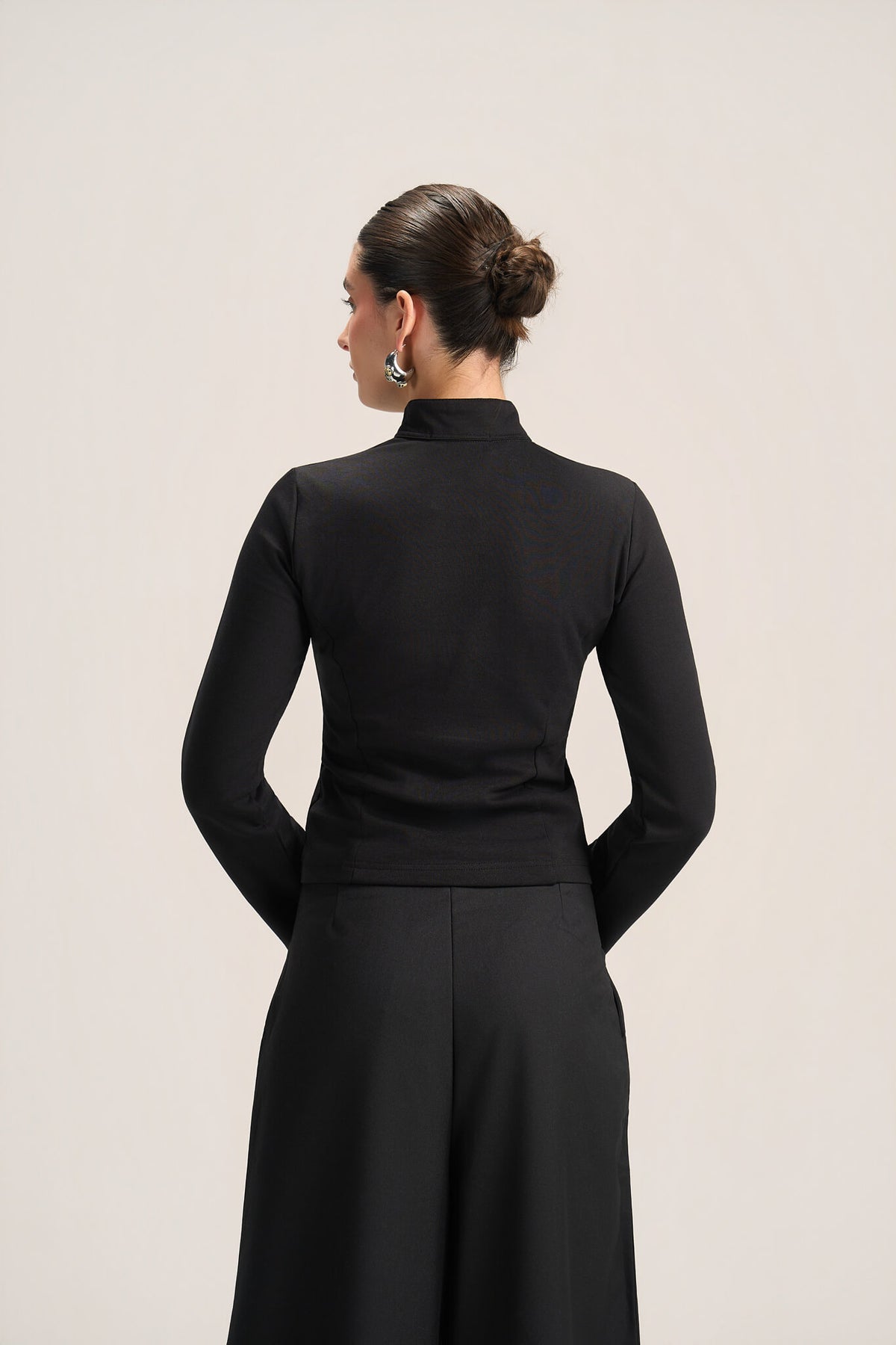 A woman shown from the back wearing a black long-sleeve mock-neck top and matching black skirt against a plain light gray studio background.