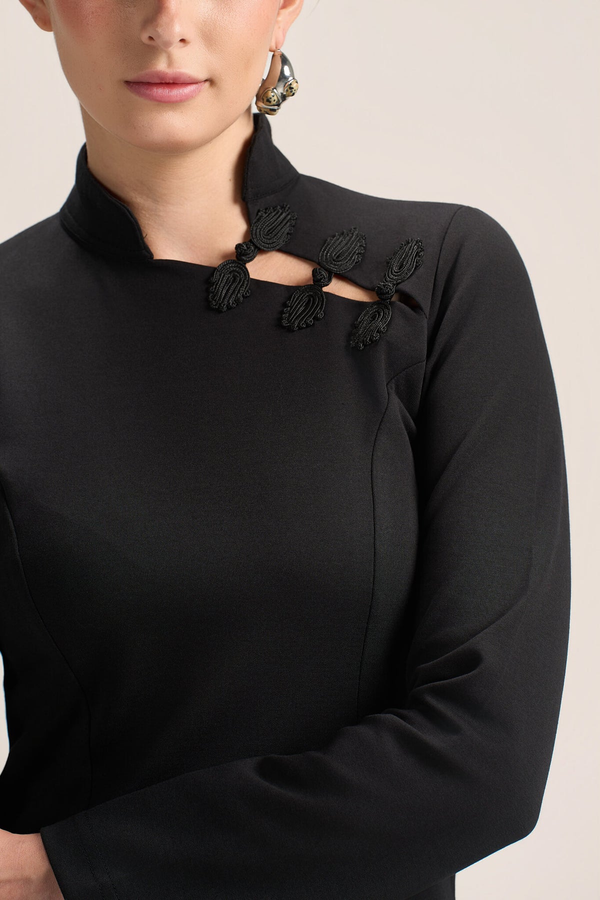 A close-up of a model wearing a black long-sleeve top with a mock neck and decorative black lace applique details across the chest.