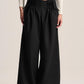 A model wearing black wide-leg pleated trousers with a gathered elastic waistband, styled with a cream ribbed cardigan and black shoes against a light beige studio background.