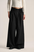 A model wearing black wide-leg pleated trousers with a gathered elastic waistband, styled with a cream ribbed cardigan and black shoes against a light beige studio background.