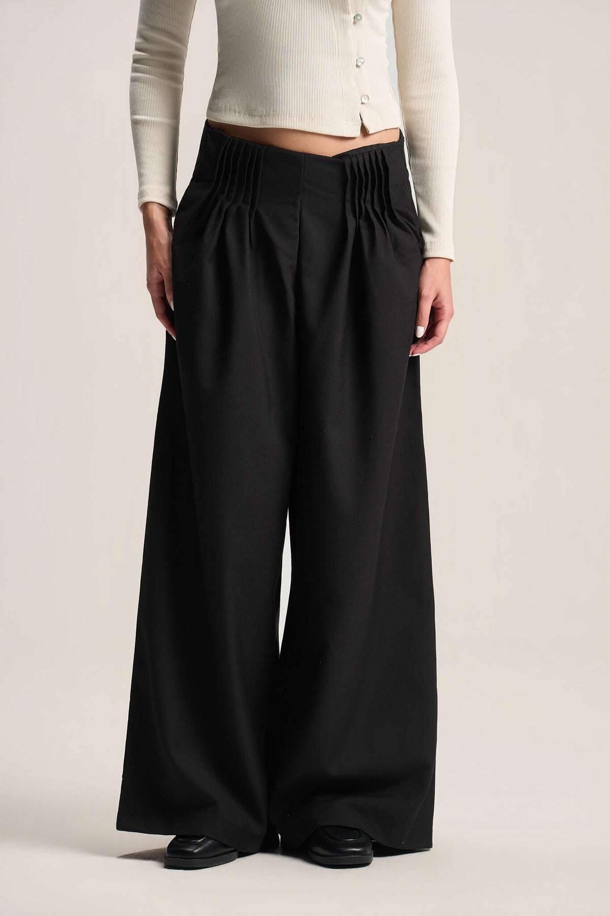 A model wearing black wide-leg pleated trousers with a gathered elastic waistband, styled with a cream ribbed cardigan and black shoes against a light beige studio background.