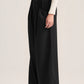 A model wearing black wide-leg pleated trousers and a cream ribbed long-sleeve top, shown from the waist down against a plain light beige studio background.