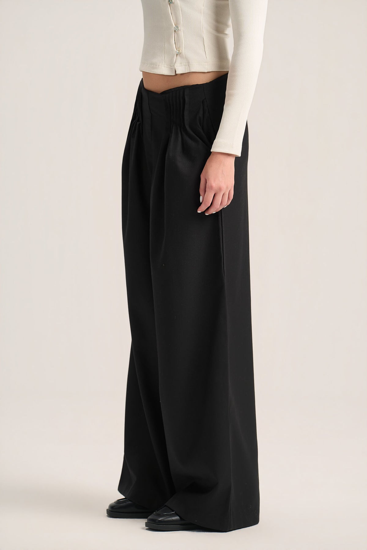 A model wearing black wide-leg pleated trousers and a cream ribbed long-sleeve top, shown from the waist down against a plain light beige studio background.