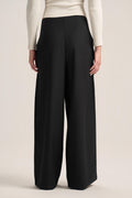 A model from the back wearing black wide-leg trousers and a cream ribbed long-sleeve top, photographed against a solid light beige studio background.