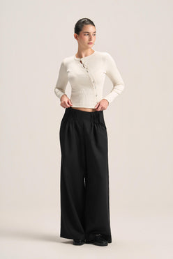 A woman modeling a cream ribbed long-sleeve cropped cardigan and black wide-leg trousers against a plain light beige studio background.