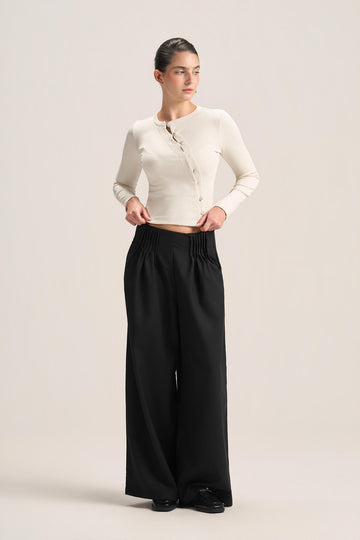 A woman modeling a cream ribbed long-sleeve cropped cardigan and black wide-leg trousers against a plain light beige studio background.