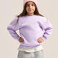A young girl modeling a light purple crewneck sweatshirt, gray sweatpants, a pink knit beanie, and white sneakers against a solid light beige background.