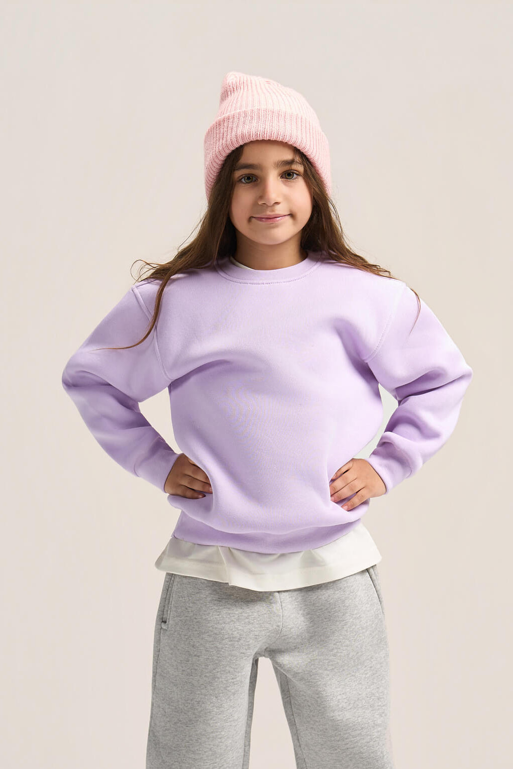 A young girl modeling a light purple crewneck sweatshirt, gray sweatpants, a pink knit beanie, and white sneakers against a solid light beige background.