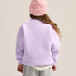 A child shown from the back wearing a light purple sweatshirt, gray sweatpants, a pink beanie, and a white undershirt, against a plain light beige studio background.