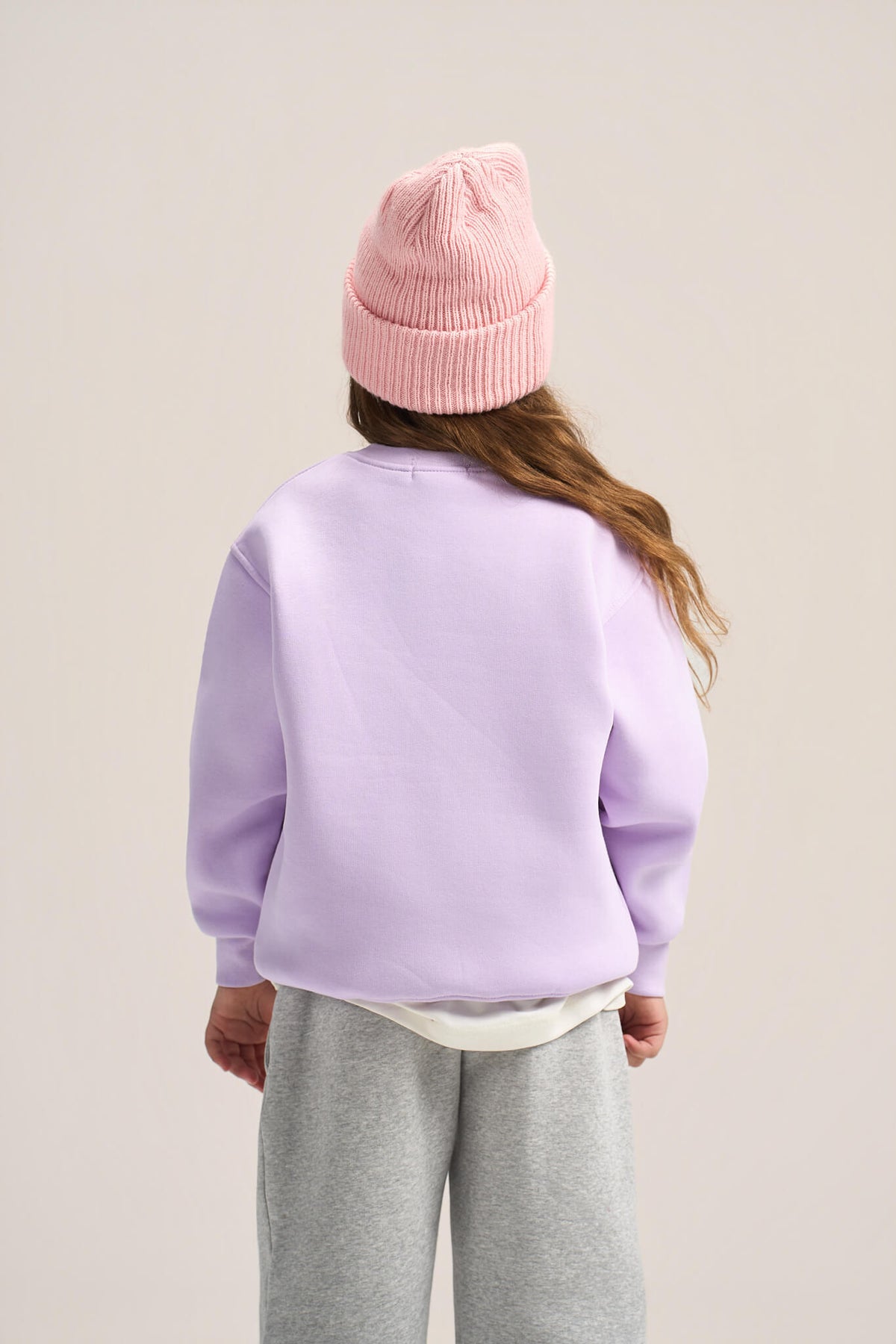 A child shown from the back wearing a light purple sweatshirt, gray sweatpants, a pink beanie, and a white undershirt, against a plain light beige studio background.