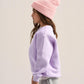 A child model shown in profile wearing a light pink ribbed knit beanie, a lavender-purple crewneck sweatshirt, and gray sweatpants against a plain light beige studio background.