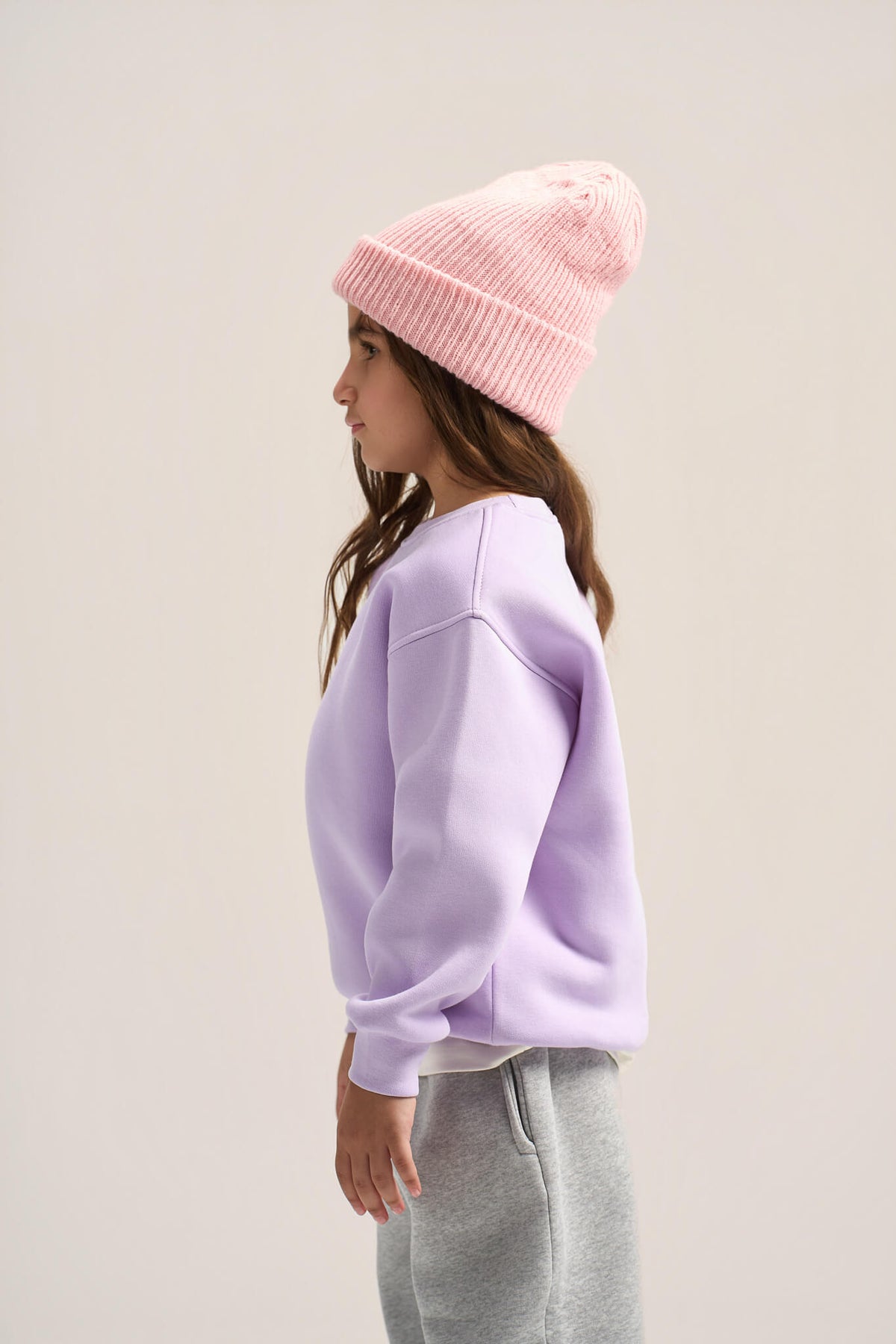 A child model shown in profile wearing a light pink ribbed knit beanie, a lavender-purple crewneck sweatshirt, and gray sweatpants against a plain light beige studio background.