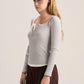 A woman modeling a light gray ribbed long-sleeve henley top with a buttoned lace-up neckline, paired with dark brown wide-leg pants, against a plain light beige studio background.