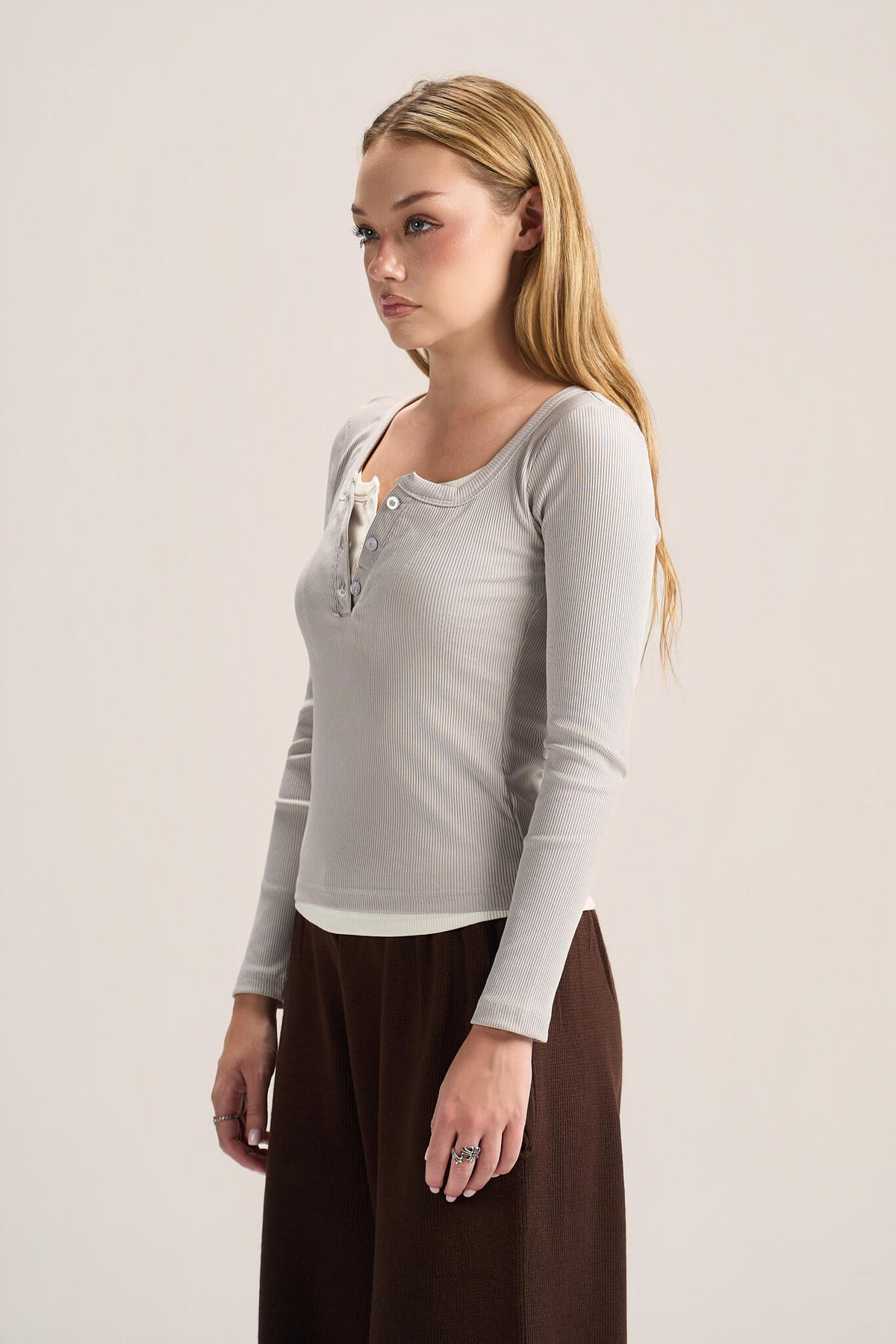 A woman modeling a light gray ribbed long-sleeve henley top with a buttoned lace-up neckline, paired with dark brown wide-leg pants, against a plain light beige studio background.