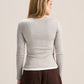 A woman viewed from the back wearing a fitted long-sleeve ribbed knit top in a light warm gray or oatmeal tone, paired with dark brown pants, against a plain light beige studio background.