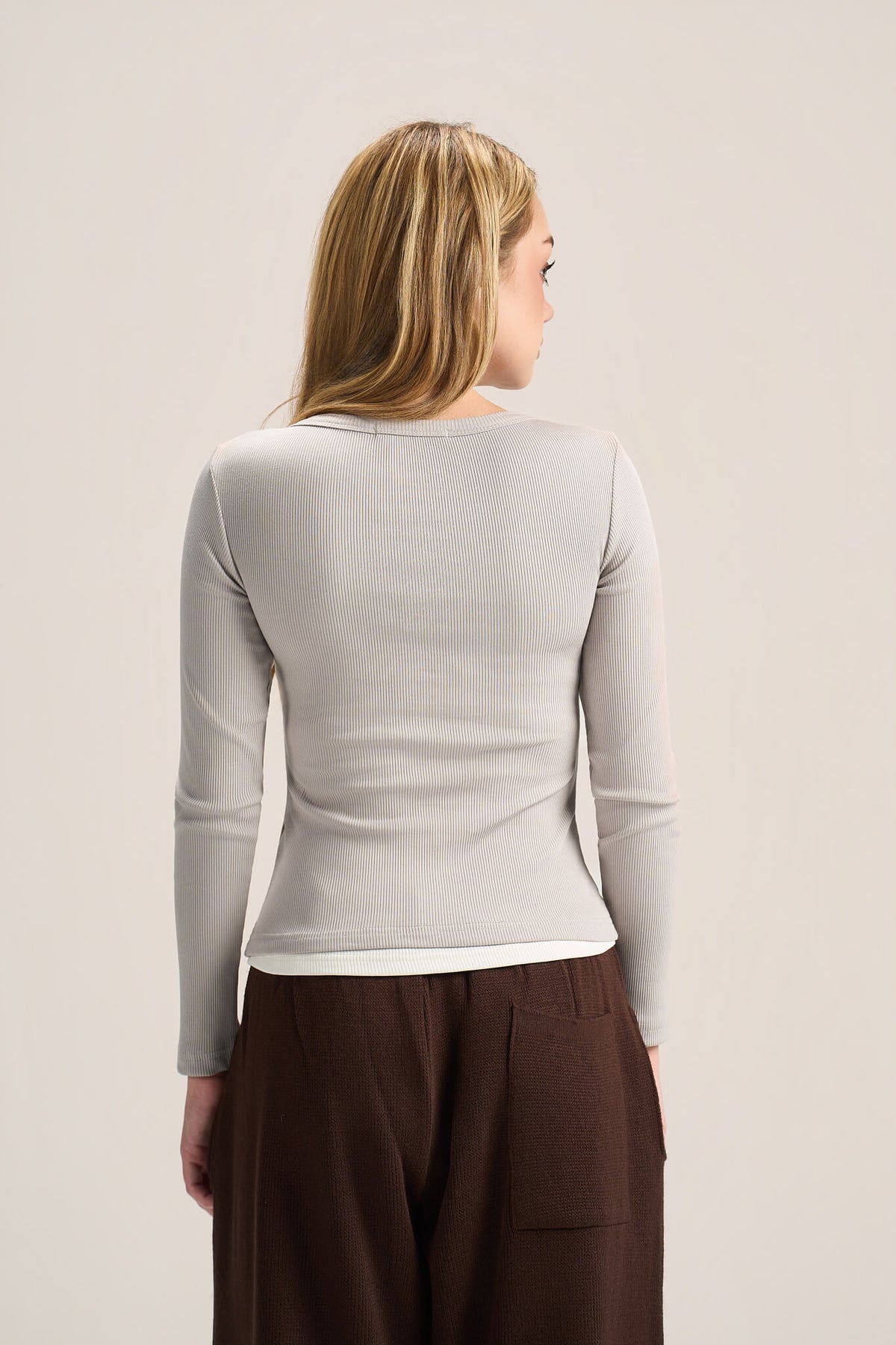 A woman viewed from the back wearing a fitted long-sleeve ribbed knit top in a light warm gray or oatmeal tone, paired with dark brown pants, against a plain light beige studio background.
