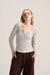 A woman modeling a light gray long-sleeve henley top with a lace-up neckline, paired with dark brown wide-leg pants, against a plain light gray studio background.