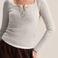 A woman wearing a light gray ribbed long-sleeve henley top with a buttoned split neckline, paired with dark brown pants, against a plain light beige background.