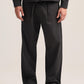 A person wearing black wide-leg trousers with pleats at the front, paired with a black jacket and black shoes, photographed against a plain light beige studio background.