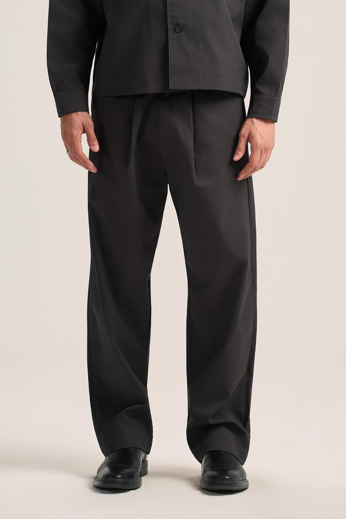 A person wearing black wide-leg trousers with pleats at the front, paired with a black jacket and black shoes, photographed against a plain light beige studio background.