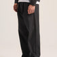 A model wearing black wide-leg trousers, a black jacket over a white t-shirt, and black leather shoes, shown from the waist down against a light beige studio background.