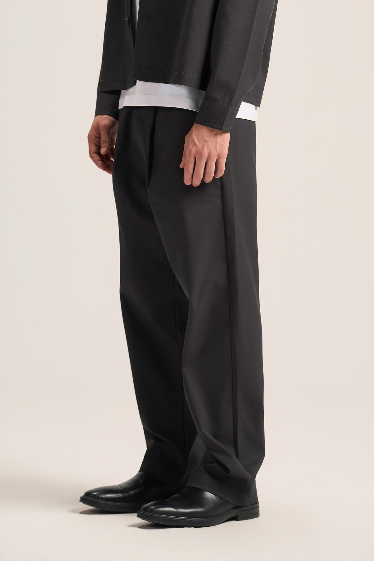 A model wearing black wide-leg trousers, a black jacket over a white t-shirt, and black leather shoes, shown from the waist down against a light beige studio background.