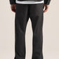 Rear view of a person wearing black wide-leg trousers, a black long-sleeve top, and black shoes against a plain light beige studio background.