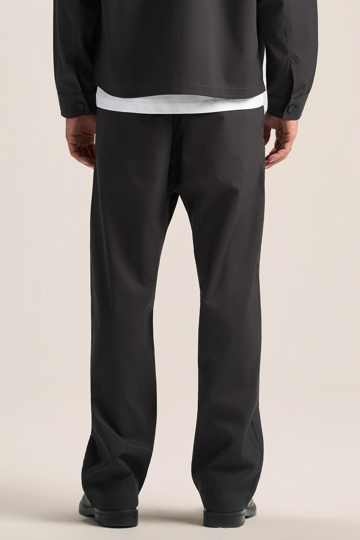 Rear view of a person wearing black wide-leg trousers, a black long-sleeve top, and black shoes against a plain light beige studio background.