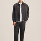 A man modeling a dark charcoal gray utility jacket over a white t-shirt, paired with dark charcoal pants and black shoes, against a solid light beige studio background.