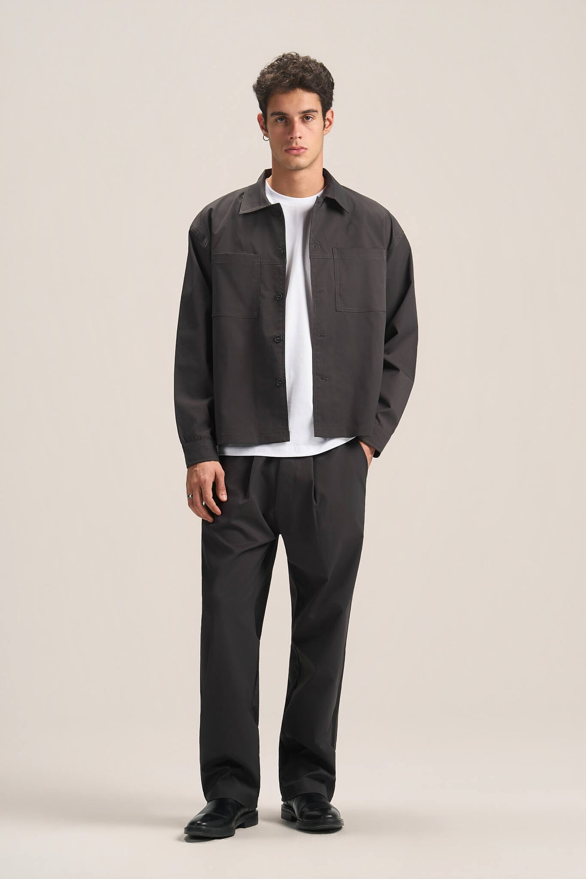 A man modeling a dark charcoal gray utility jacket over a white t-shirt, paired with dark charcoal pants and black shoes, against a solid light beige studio background.