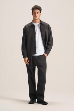 A man modeling a dark charcoal gray overshirt, black trousers, and a white t-shirt against a solid light beige studio background.