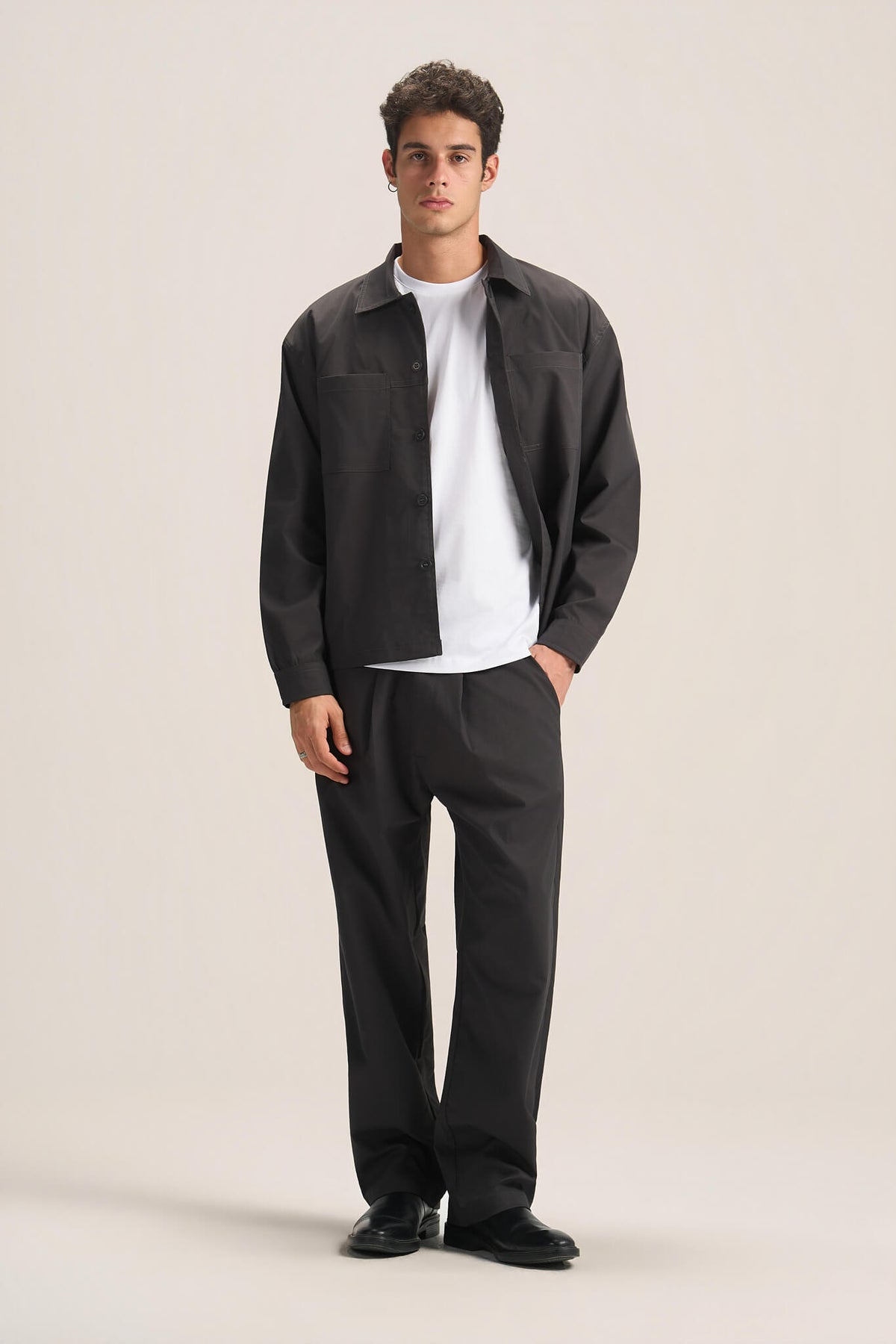 A man modeling a dark charcoal gray overshirt, black trousers, and a white t-shirt against a solid light beige studio background.
