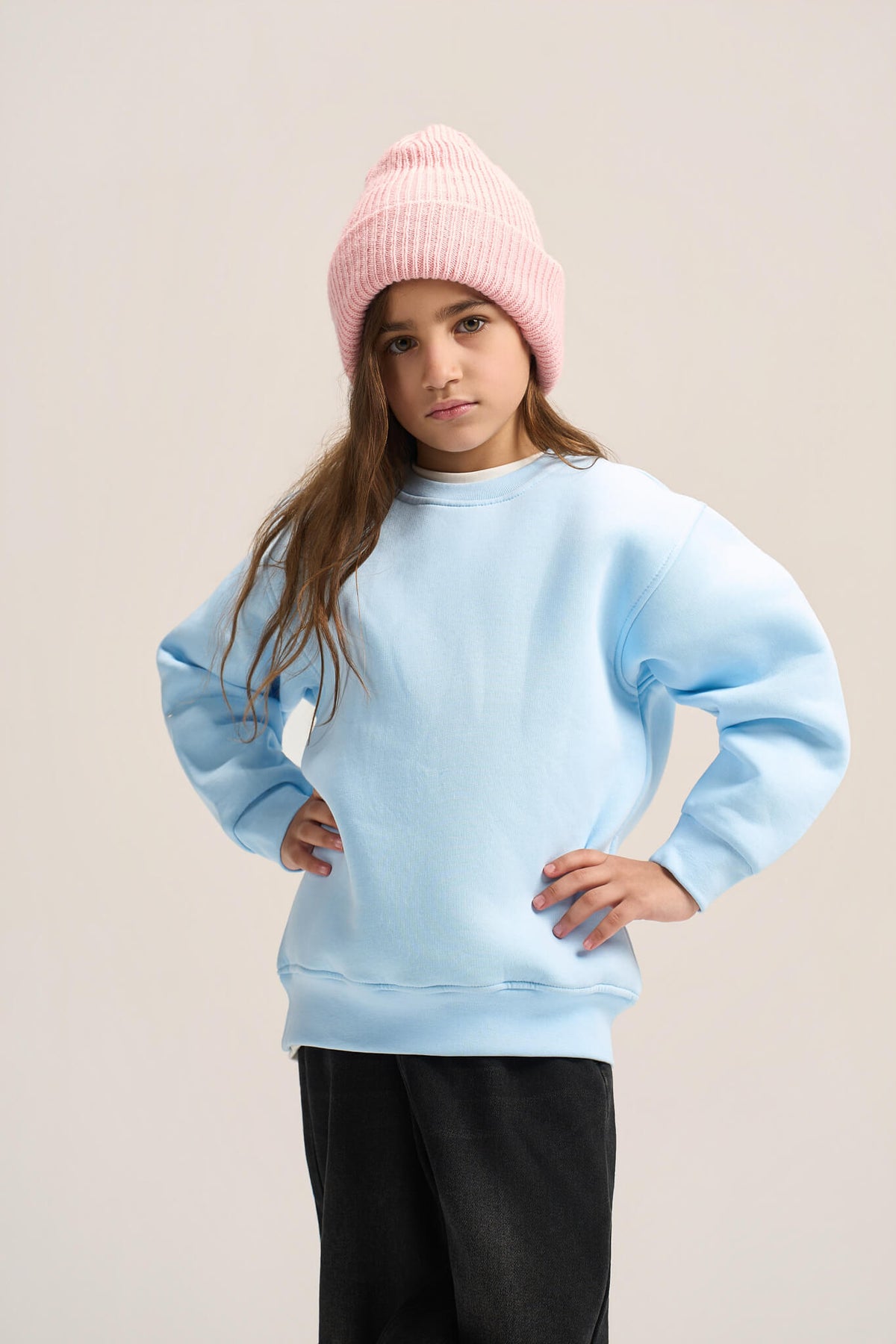 A young girl wearing a light blue crewneck sweatshirt, a pink ribbed knit beanie, and black pants, posing against a solid light beige background.