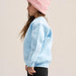 A child shown in side profile wearing a light pink ribbed knit beanie, a light blue sweatshirt, and black pants against a plain light beige studio background.