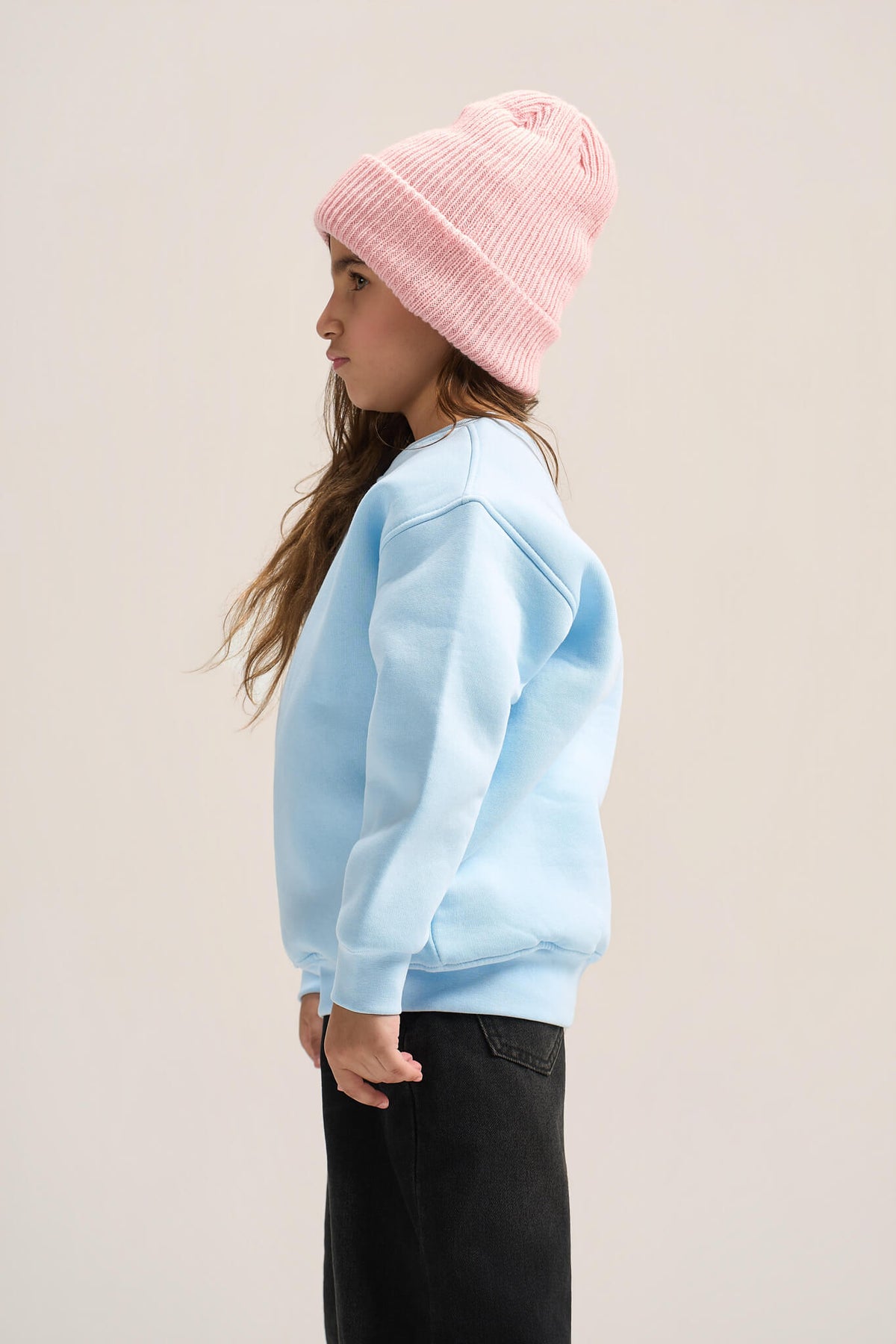 A child shown in side profile wearing a light pink ribbed knit beanie, a light blue sweatshirt, and black pants against a plain light beige studio background.