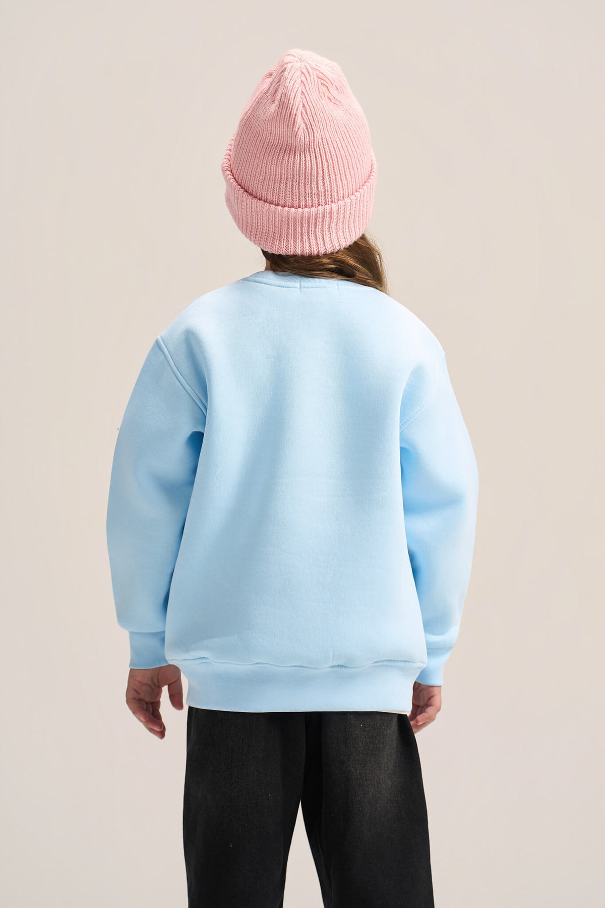 A child shown from the back wearing a light blue crewneck sweatshirt, a pink ribbed knit beanie, and black pants, set against a plain light beige studio background.