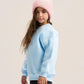 A young girl wearing a light blue crewneck sweatshirt, a pink ribbed knit beanie, and black pants, standing against a solid light beige studio background.