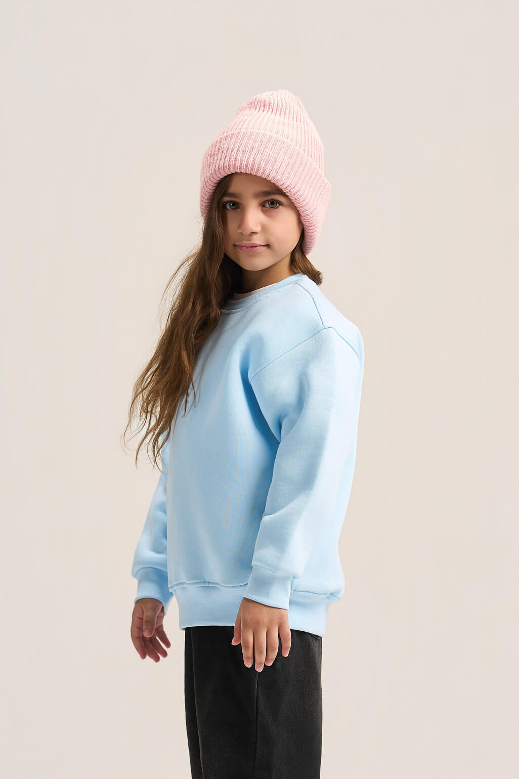 A young girl wearing a light blue crewneck sweatshirt, a pink ribbed knit beanie, and black pants, standing against a solid light beige studio background.
